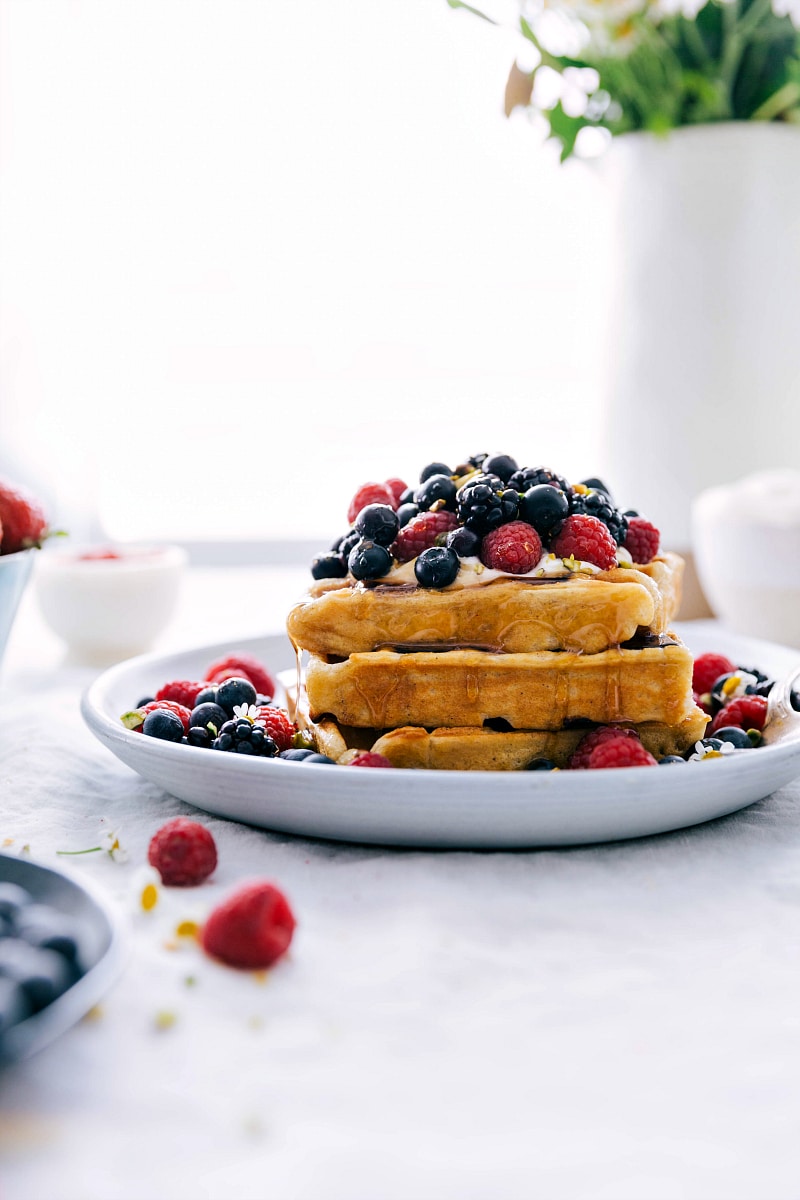 Image of a stack of waffles with berries and whipped cream on top with syrup drizzling down.